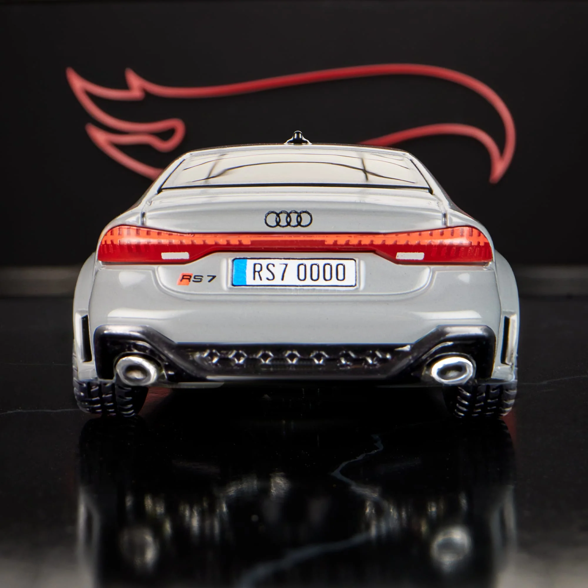 Model samochodu w kolorze szarym z widocznym logo Audi i tablicą rejestracyjną \'RS7 0000\'. W tle znajduje się czerwony symbol przypominający płomień.