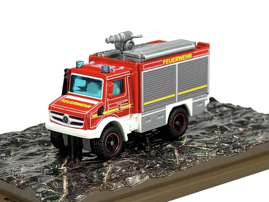 Model metalowego wozu strażackiego w kolorze czerwonym z napisem \'FEUERWEHR\' na boku, umieszczony na podstawce imitującej teren. Na przodzie pojazdu widoczny jest napis \'FEUERWEHR\' oraz logo producenta.