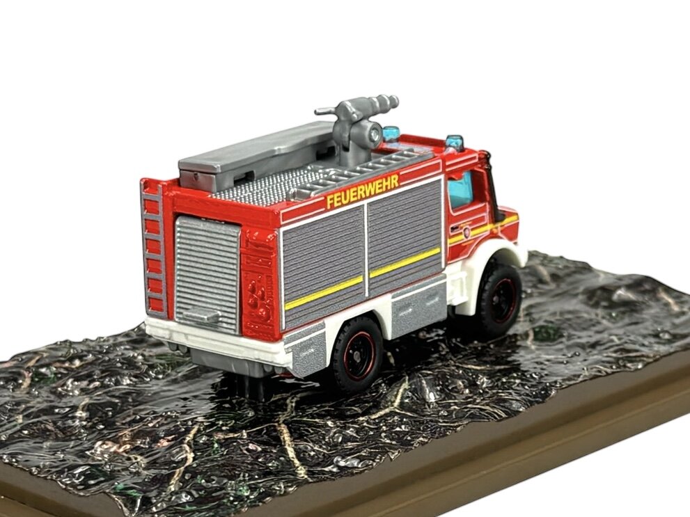 Model metalowego wozu strażackiego z napisem \'FEUERWEHR\' na boku, umieszczony na podstawce imitującej teren. Pojazd jest w kolorze czerwonym z szarymi i białymi elementami.