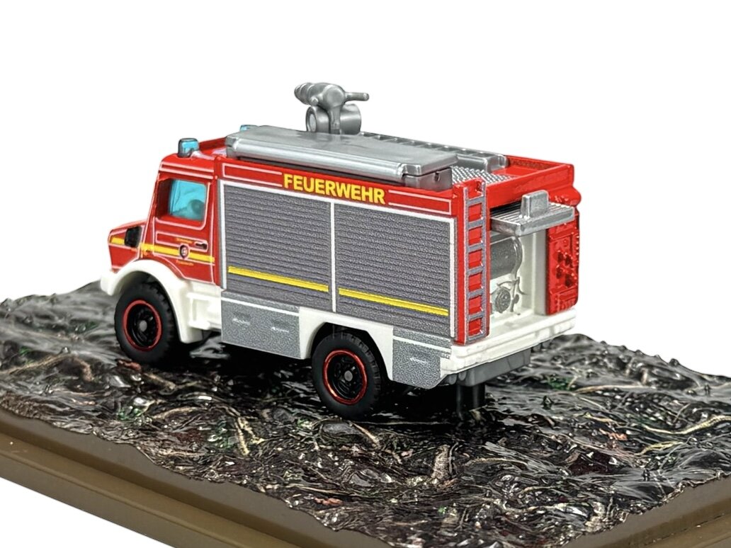 Model metalowego wozu strażackiego w kolorze czerwonym z napisem \'FEUERWEHR\' na boku, umieszczony na podstawce imitującej teren. Pojazd posiada szczegółowe elementy, takie jak drabina i reflektory.