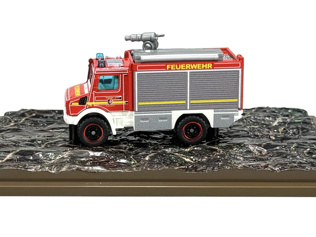 Model metalowego wozu strażackiego w czerwono-białych barwach z napisem \'FEUERWEHR\' na boku, umieszczony na podstawce imitującej nierówną powierzchnię.