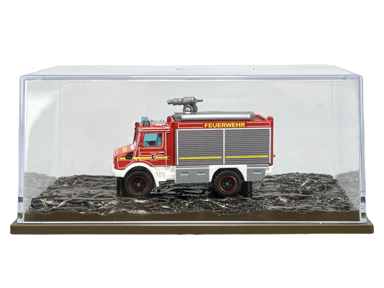 Model metalowego wozu strażackiego Unimog w czerwonym kolorze z napisem \'FEUERWEHR\' na boku, umieszczony w przezroczystej gablocie na czarnej podstawce.
