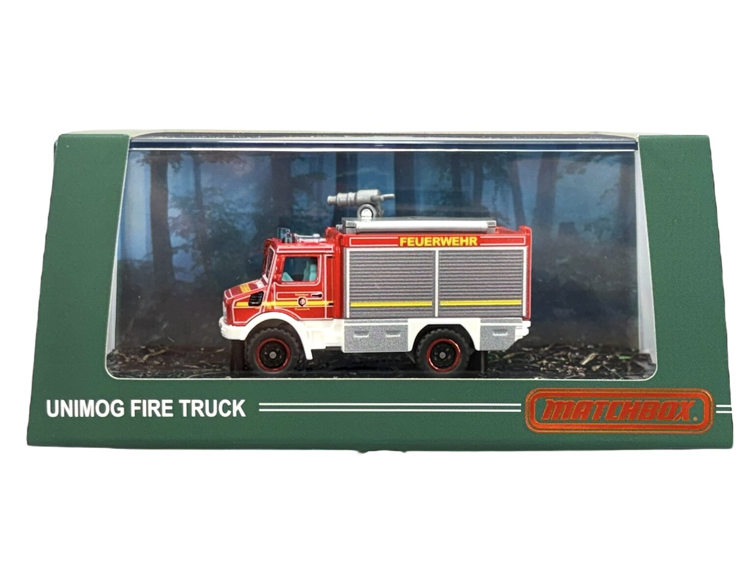 Model metalowego wozu strażackiego Unimog w czerwonym kolorze z napisem \'FEUERWEHR\' na boku, zapakowany w zielone pudełko z przezroczystym frontem. Na pudełku widnieje napis \'UNIMOG FIRE TRUCK\' oraz logo \'matchbox\'.