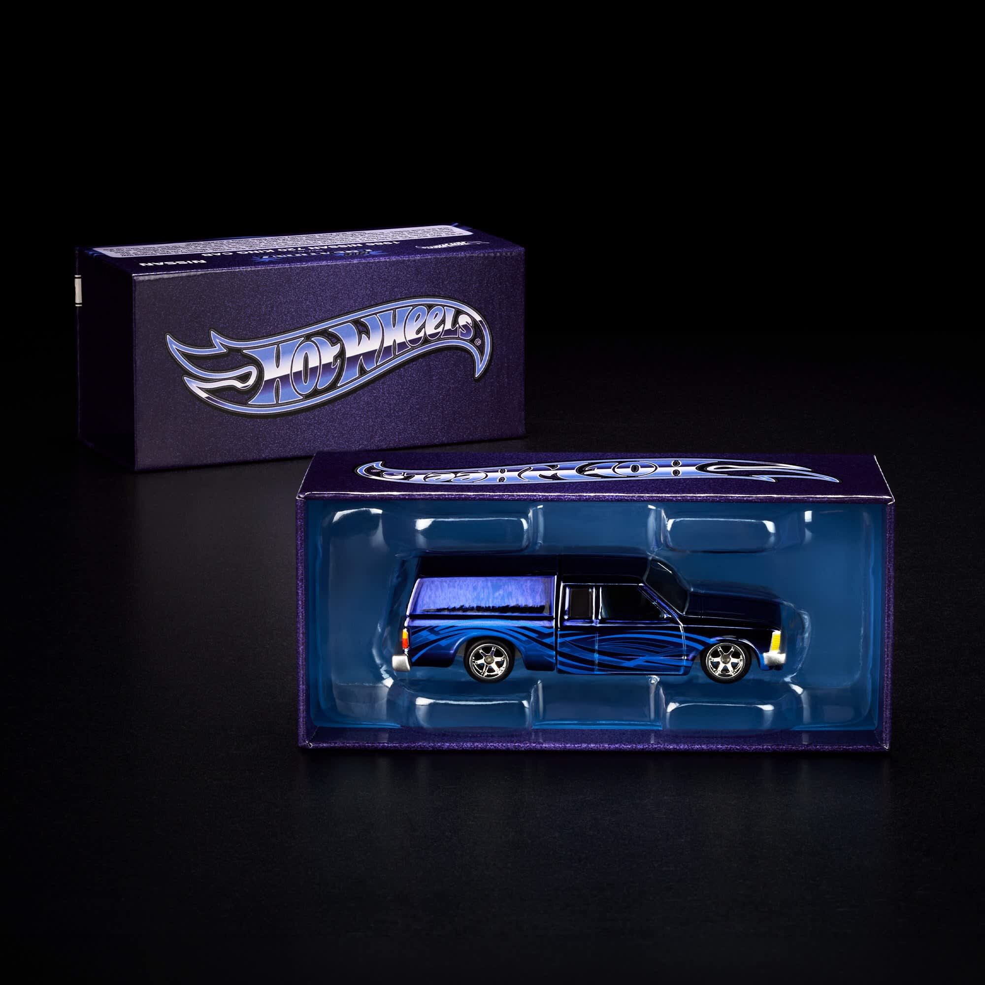 Na obrazie widoczny jest model samochodu w kolorze niebieskim z czarnymi i niebieskimi wzorami, umieszczony w przezroczystym opakowaniu. W tle znajduje się pudełko z logo \'Hot Wheels\'.