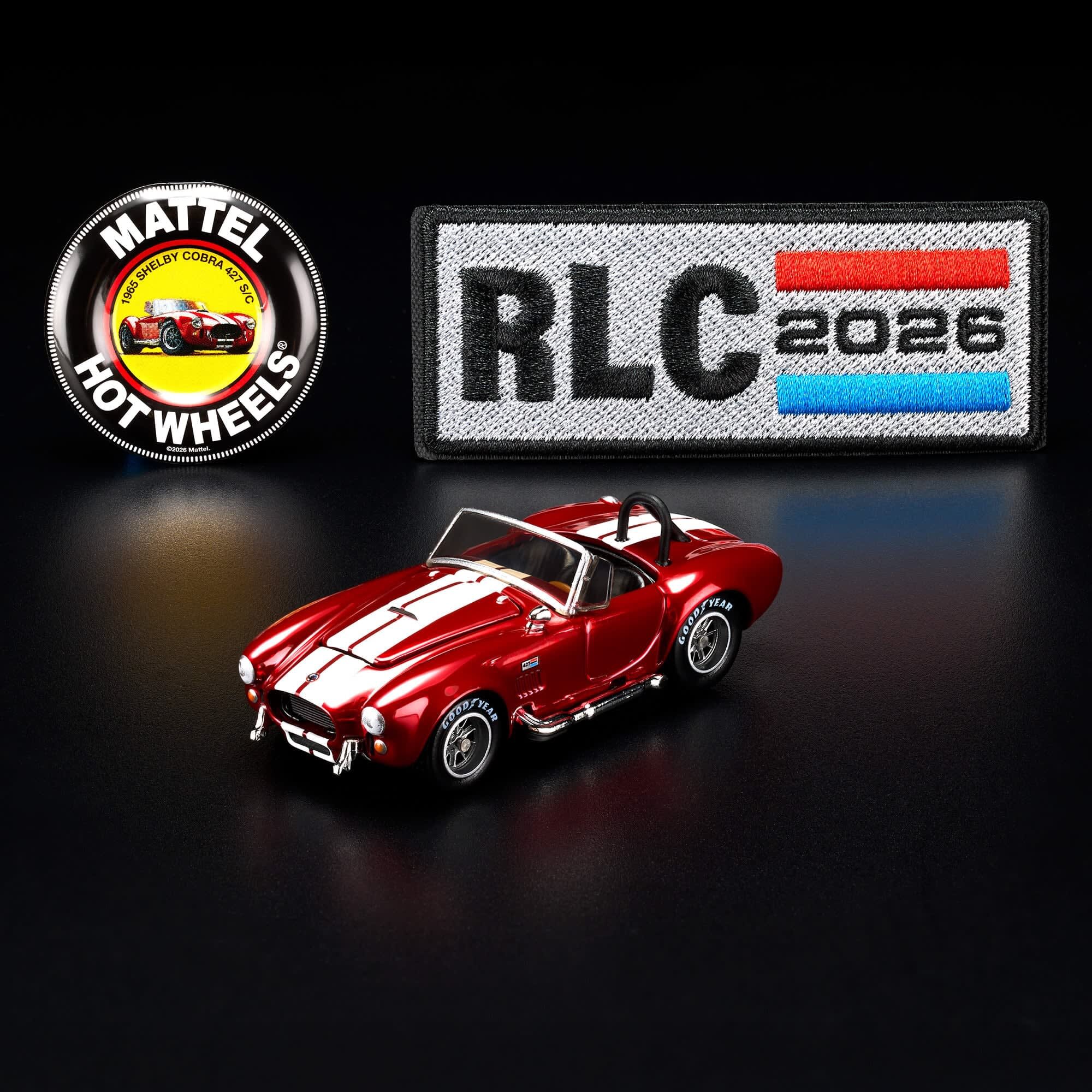 Na obrazie znajduje się czerwony model samochodu z białymi pasami, obok którego widoczne są dwa emblematy: okrągły z napisem \'MATTEL HOT WHEELS\' oraz prostokątny z napisem \'RLC 2026\'.