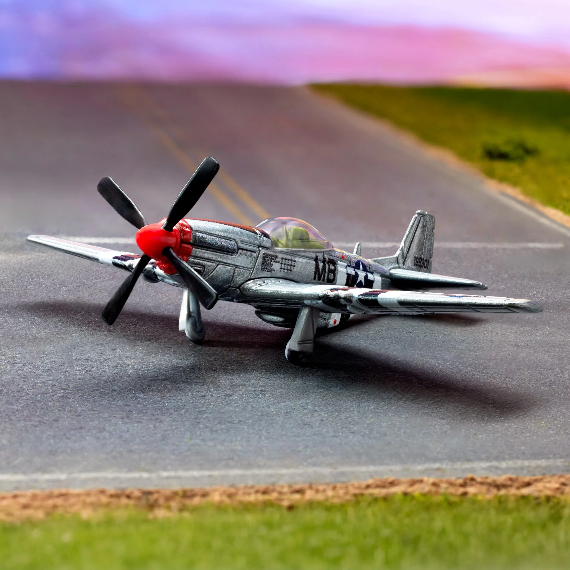 Model metalowego samolotu P-51 Mustang z czerwonym nosem i czarnymi śmigłami stoi na drodze. Na kadłubie widoczne są oznaczenia \'MB\' oraz wzór w kształcie gwiazdy.