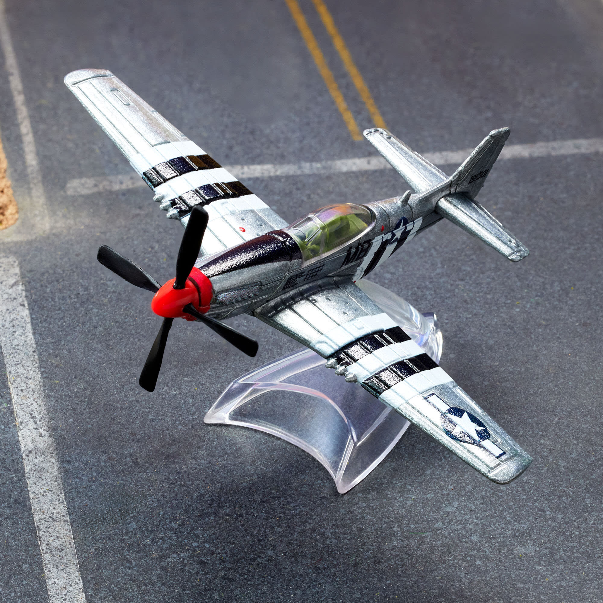Model metalowego samolotu P-51 Mustang w kolorze srebrnym z czarnymi i białymi pasami, umieszczony na przezroczystej podstawce. Samolot ma czerwony nos i czarny śmigło, a w tle widoczna jest powierzchnia przypominająca pas startowy.
