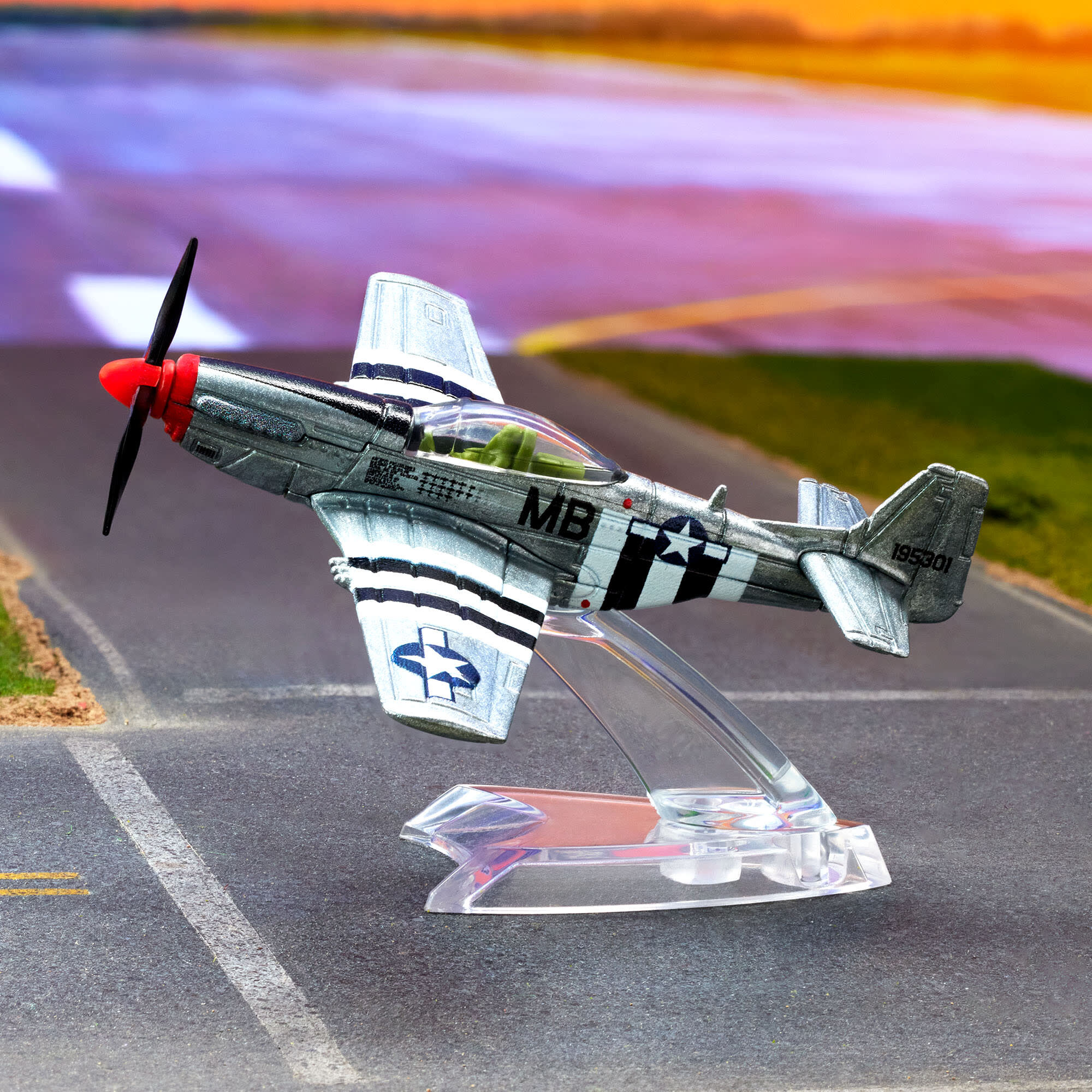 Model metalowego samolotu P-51 Mustang na przezroczystej podstawce, z widocznymi oznaczeniami \'MB\' i \'19530\'. W tle rozmyta sceneria przypominająca pas startowy.