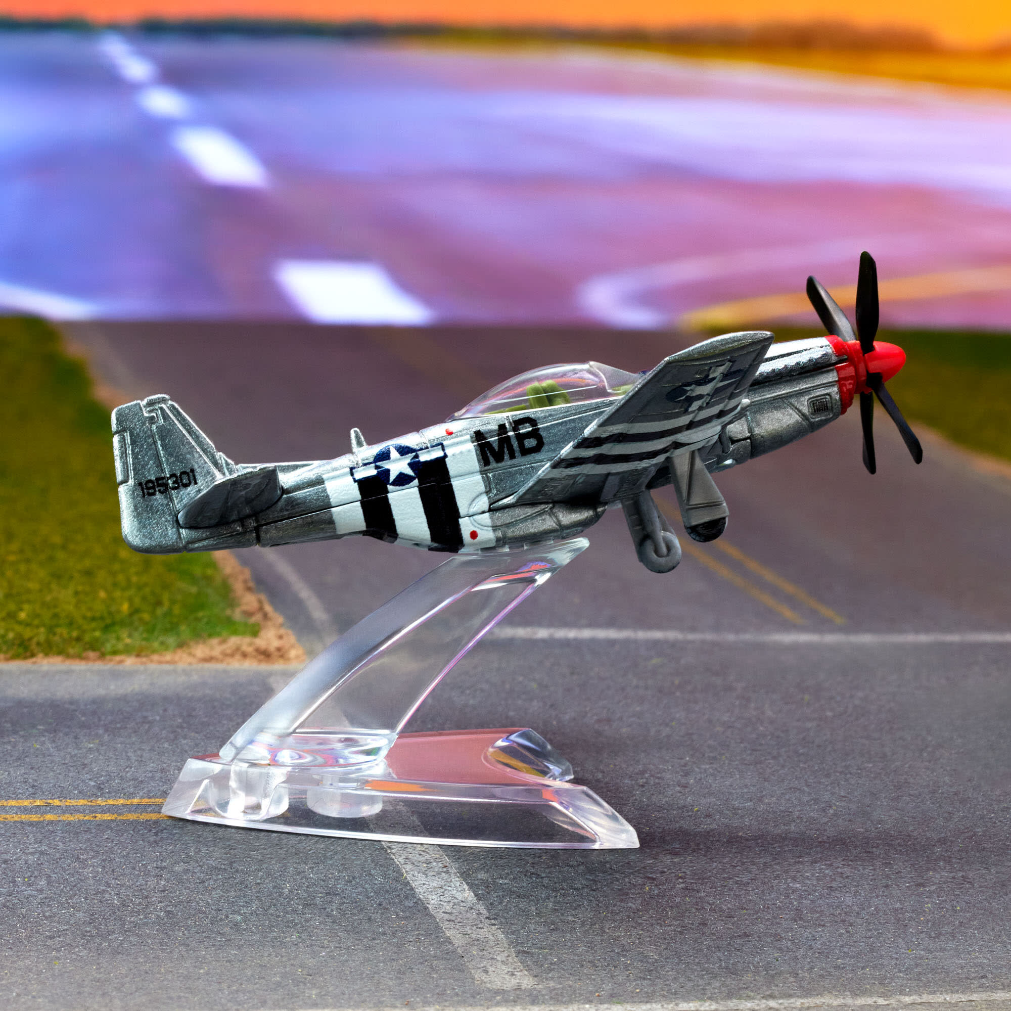 Model samolotu P-51 Mustang w kolorze srebrnym z czarnymi i białymi pasami, umieszczony na przezroczystej podstawce. Na kadłubie widoczne są oznaczenia \'MB\' i \'195301\'.