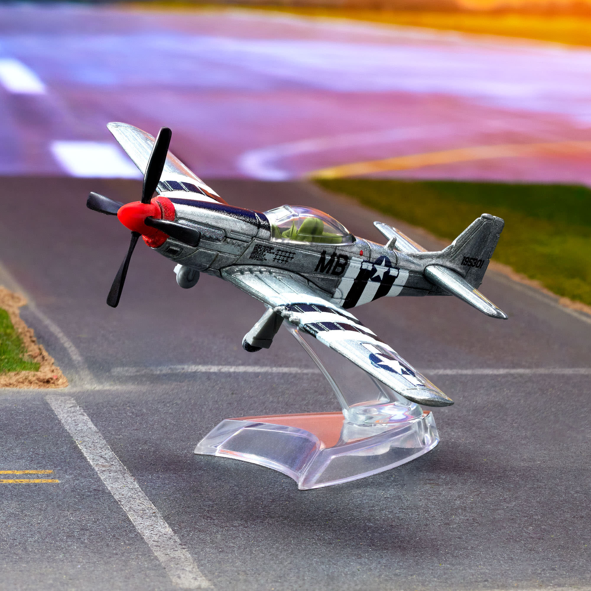 Model metalowego samolotu P-51 Mustang na przezroczystej podstawce, z widocznymi oznaczeniami \'MB\' i czarnymi pasami na skrzydłach. Tło przedstawia rozmyty obraz pasa startowego.