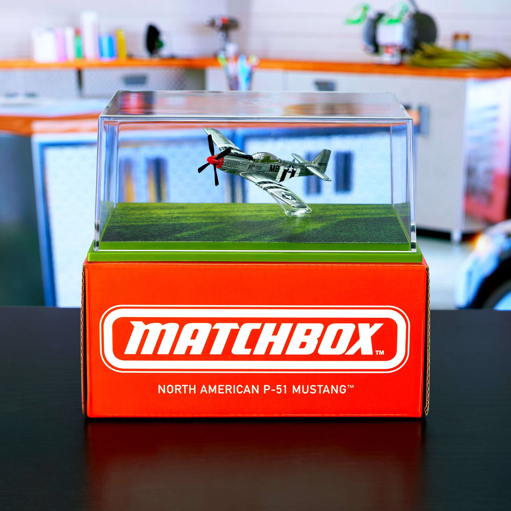 Model samolotu w przezroczystej gablocie stoi na czerwonym pudełku z napisem \'MATCHBOX NORTH AMERICAN P-51 MUSTANG\'. W tle widoczne są rozmyte elementy wnętrza, takie jak półki i przedmioty biurowe.