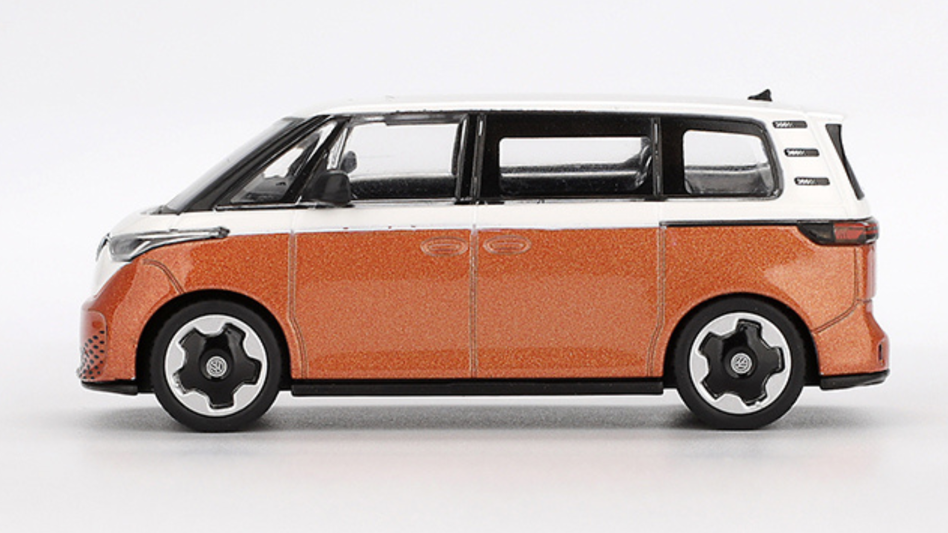 Model samochodu w skali 1:64 przedstawia pomarańczowy pojazd z białym dachem i czarnymi szybami. Koła mają czarno-białe felgi z widocznym logo VW.