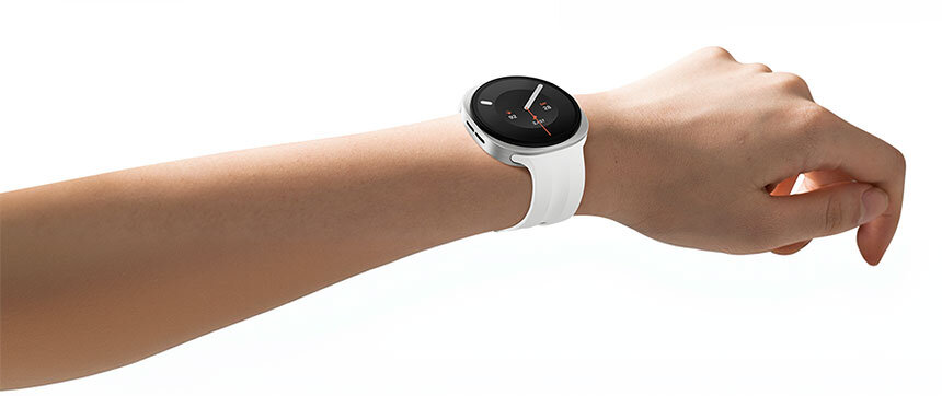 Na zdjęciu widoczny jest smartwatch z okrągłym wyświetlaczem i białym paskiem, założony na nadgarstku. Ekran zegarka pokazuje prosty interfejs z białymi wskazówkami na czarnym tle.