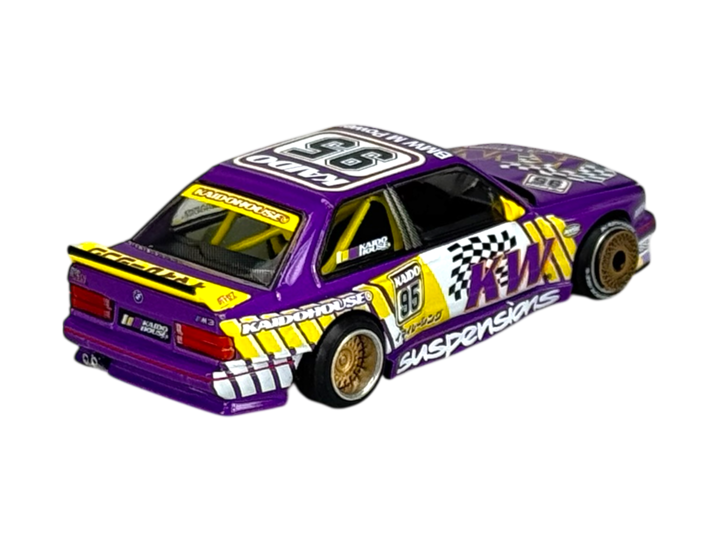 Model samochodu w skali 1:64, przedstawiający BMW M3 E30, w kolorze fioletowym z żółtymi i białymi akcentami oraz licznymi naklejkami, w tym \'KAIDO\', \'KW\', \'SUSPENSIONS\'.