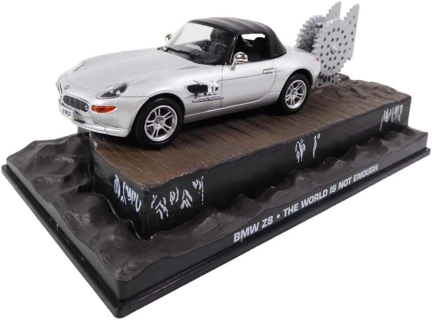 Model samochodu BMW Z8 w kolorze srebrnym z czarnym dachem, umieszczony na podstawce z napisem \'BMW Z8 - THE WORLD IS NOT ENOUGH\'. W tle widoczny jest duży, srebrny element przypominający piłę tarczową.