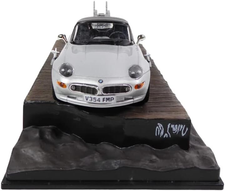 Model samochodu w skali 1:43 przedstawiający biały pojazd z tablicą rejestracyjną V354 FMP, umieszczony na podstawce z imitacją terenu. Na podstawce widoczne są nieczytelne napisy.