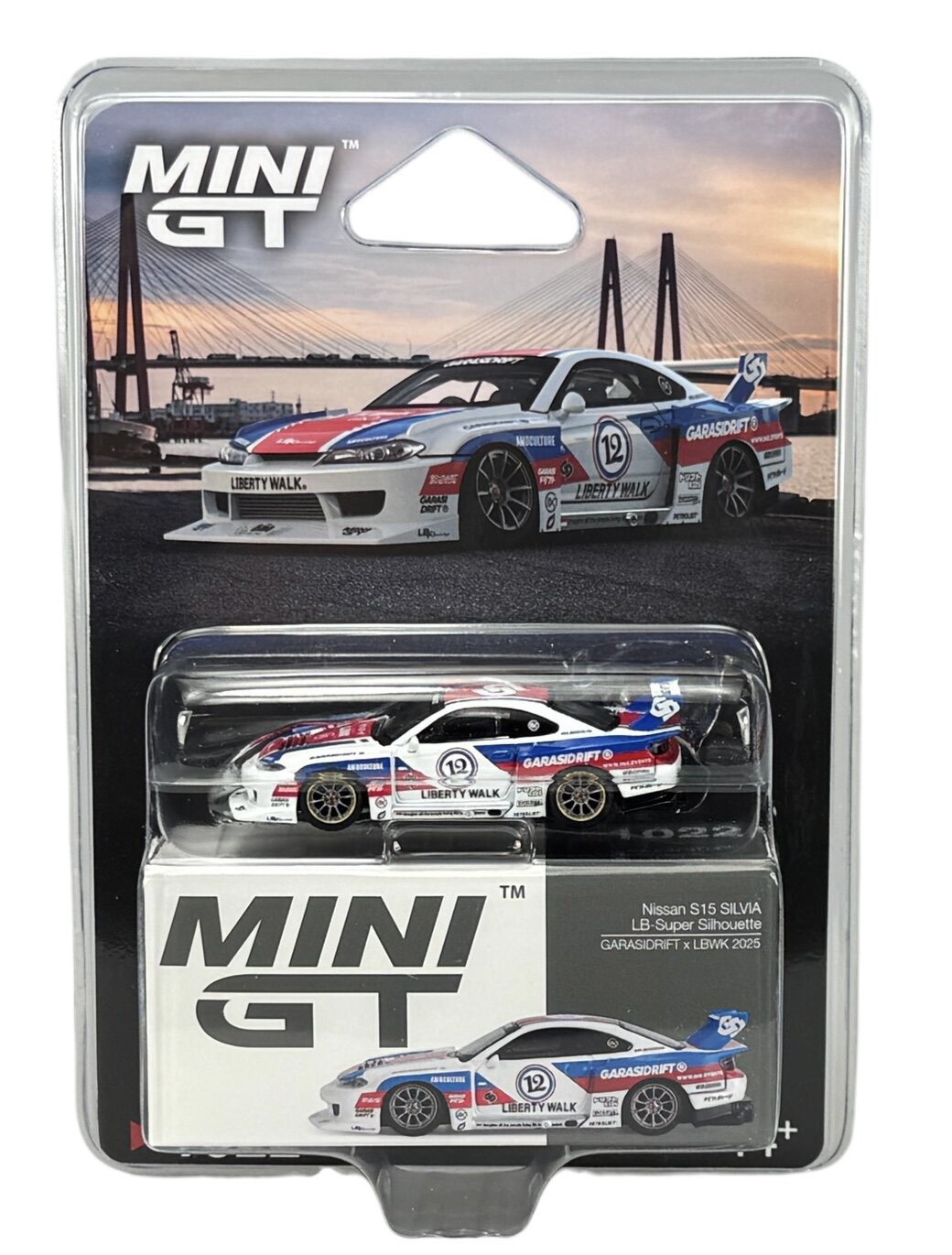 Opakowanie modelu samochodu Mini GT z widocznym modelem Nissan S15 Silvia w kolorach biało-czerwono-niebieskich. Na opakowaniu znajduje się napis \'MINI GT\' oraz zdjęcie pełnowymiarowego samochodu na tle mostu.