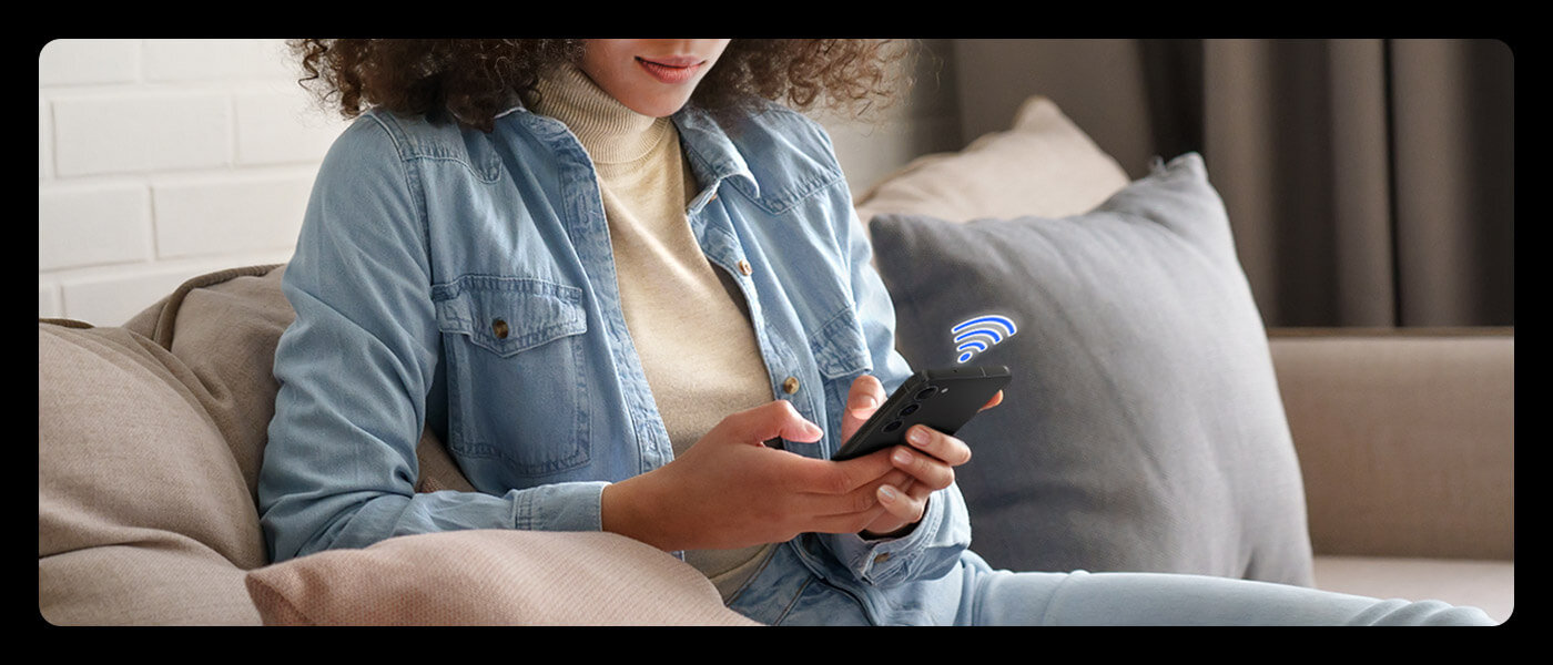 Skorzystaj z aplikacji SmartThings, by zarządzać urządzeniami Samsung z dowolnego miejsca Osoba siedzi na kanapie, trzymając w rękach smartfon. Obok telefonu widoczna jest ikona sygnału bezprzewodowego.