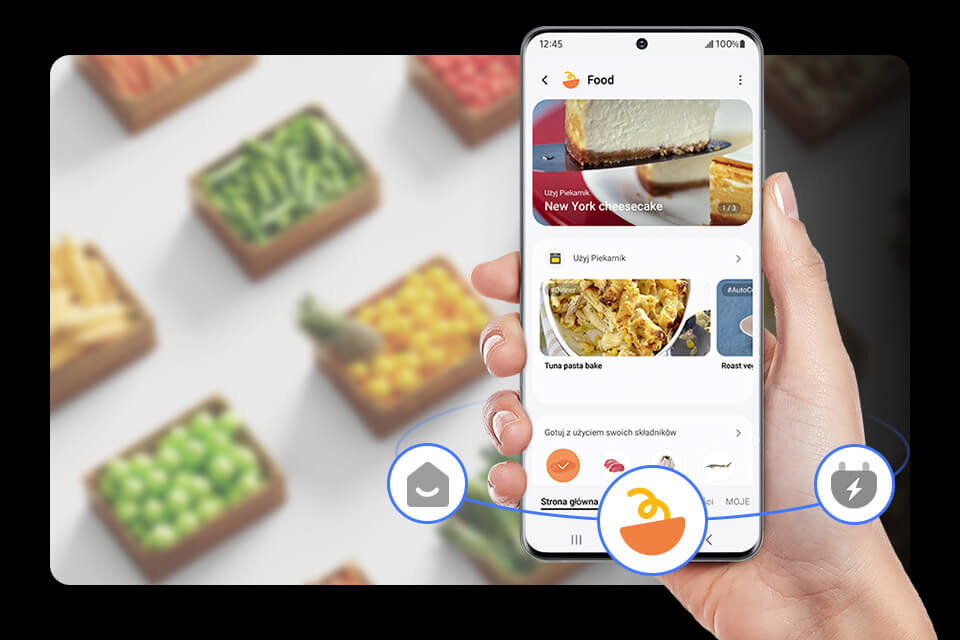 smart things food 960 Osoba trzyma smartfon z aplikacją kulinarną, na ekranie widoczny jest przepis na \'New York cheesecake\'. W tle rozmyte pudełka z warzywami.