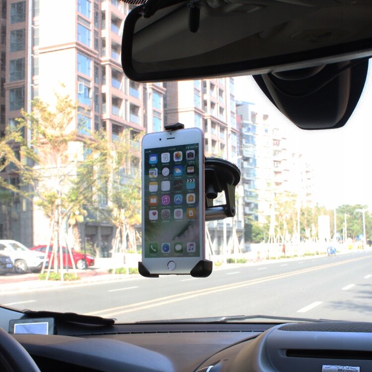 Wnętrze samochodu z zamontowanym na szybie uchwytem samochodowym, w którym znajduje się telefon z widocznym ekranem głównym. W tle widoczne są budynki i drzewa za szybą samochodu.