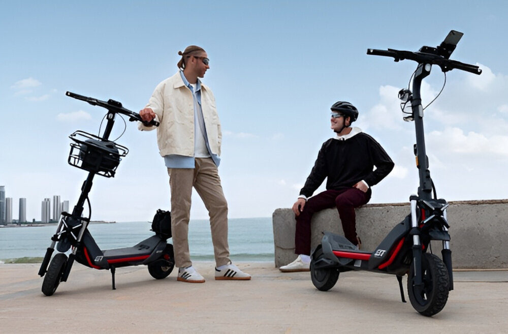 Na obrazie widać dwie osoby stojące obok dwóch czarnych hulajnóg elektrycznych z logo \'SEGWAY\'. W tle znajduje się morze i kilka wysokich budynków.