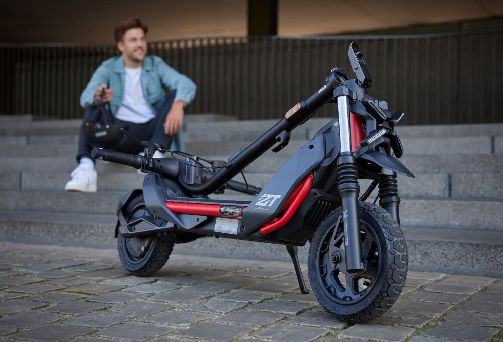 Na zdjęciu znajduje się czarna hulajnoga elektryczna z czerwonymi akcentami, ustawiona na chodniku. W tle widać osobę siedzącą na schodach.