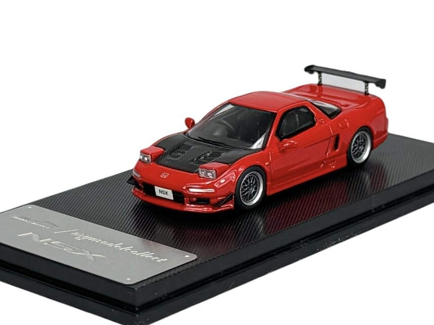 Czerwony model samochodu w skali 1:64 z czarnymi elementami na masce i dużym tylnym spojlerem, umieszczony na czarnej podstawce. Na podstawce widoczny jest napis \'NSX\'.