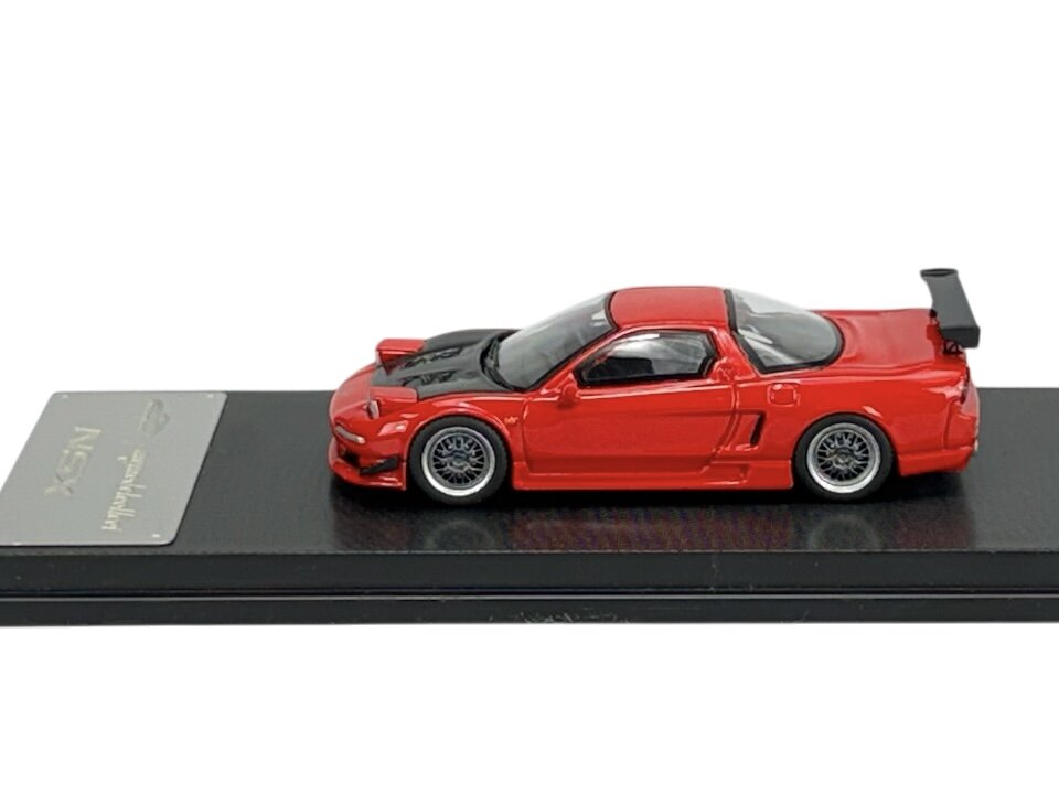 Czerwony model samochodu sportowego z czarnym dachem i dużym tylnym spojlerem, umieszczony na czarnej podstawce. Na podstawce widoczna jest metalowa tabliczka z napisem \'NSX\'.