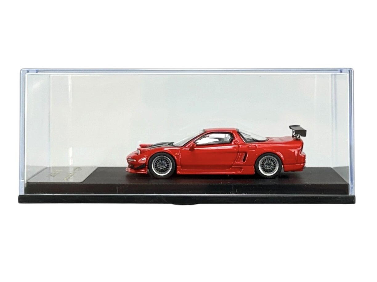 Czerwony model samochodu w skali 1:64 umieszczony w przezroczystej gablocie na czarnej podstawce. Samochód ma widoczny duży tylny spojler i szczegółowe felgi.