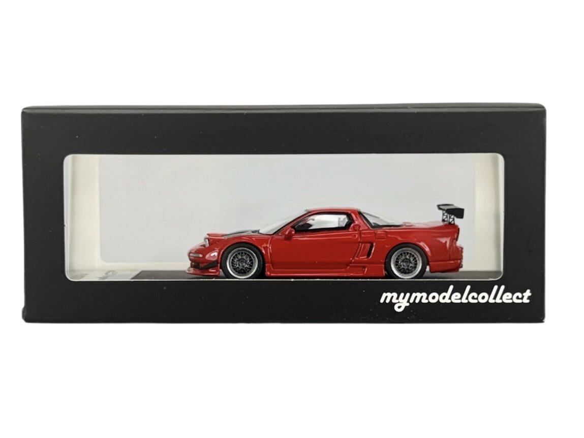 Model czerwonego samochodu w skali 1:64 znajduje się w czarnym pudełku z przezroczystym frontem. Na pudełku widnieje napis \'mymodelcollect\'.