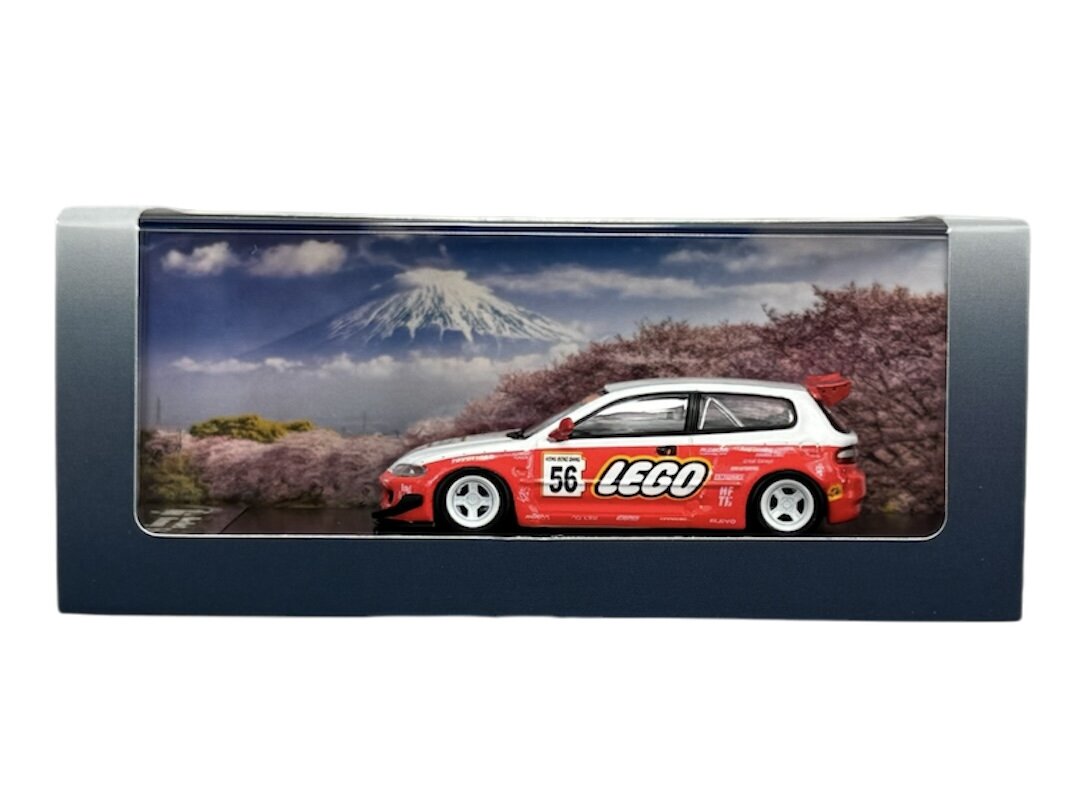 Model samochodu w skali 1:64 w czerwonym kolorze z napisem \'LEGO\' na boku, umieszczony w pudełku z tłem przedstawiającym górski krajobraz. Na samochodzie widoczny jest numer 56.