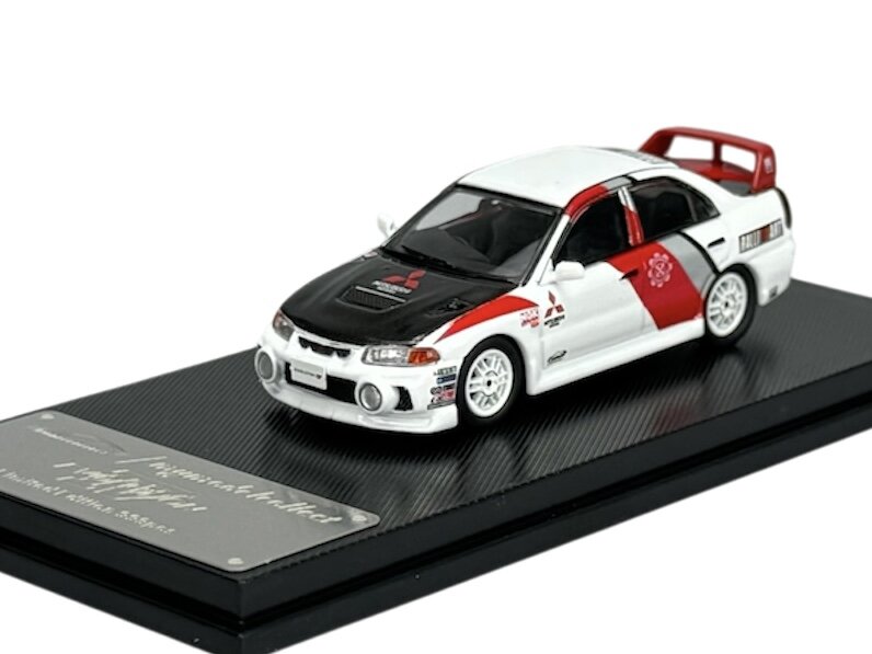 Model samochodu w skali 1:64 przedstawiający biały Mitsubishi Lancer Evolution z czarnymi i czerwonymi akcentami, umieszczony na czarnej podstawce z metalową tabliczką. Na masce widoczne jest logo Mitsubishi.