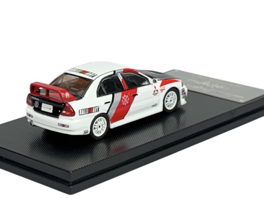 Model samochodu w skali 1:64 przedstawia biały pojazd z czerwonymi i czarnymi detalami, umieszczony na czarnej podstawce. Na samochodzie widoczne są napisy \'RALLI ART\' oraz logo producenta.