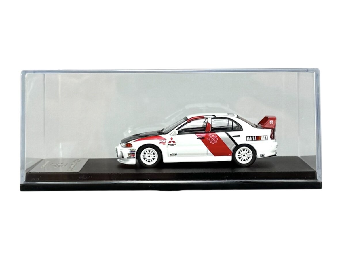 Model samochodu w skali 1:64 przedstawiający biały Mitsubishi Lancer Evolution IV z czerwonymi i czarnymi detalami, umieszczony w przezroczystej gablocie na czarnej podstawce.