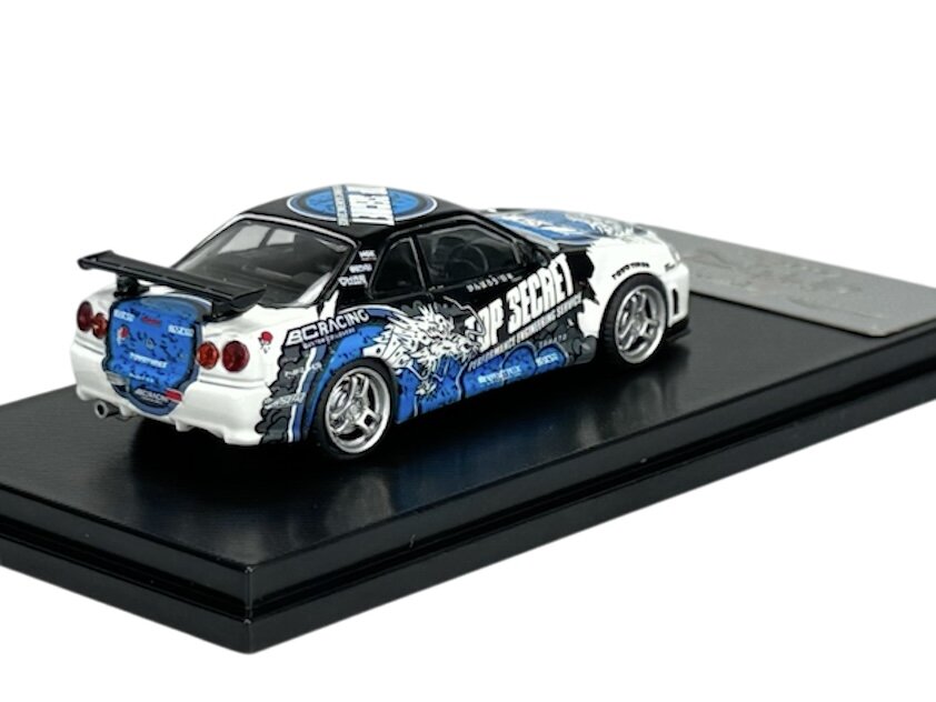 Model samochodu w skali 1:64 przedstawia sportowy pojazd z napisami \'TOP SECRET\' i grafiką w kolorach niebieskim, białym i czarnym. Auto umieszczone jest na czarnej podstawce.