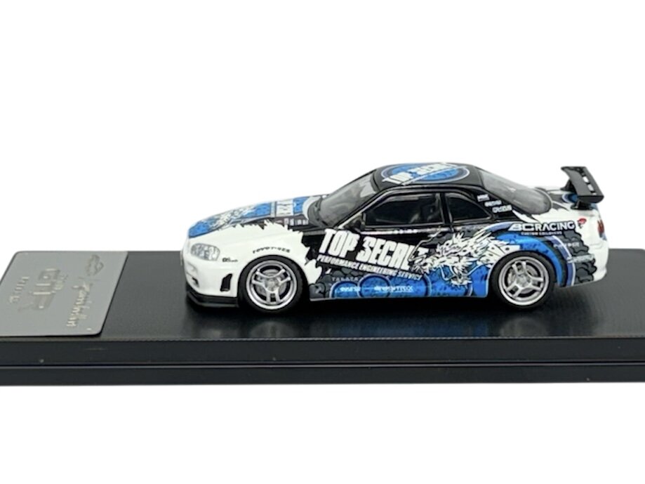 Model samochodu w skali 1:64 przedstawia sportowy pojazd z napisami \'TOP SECRET\' i grafiką w kolorach białym, czarnym i niebieskim. Samochód umieszczony jest na czarnej podstawce z metalową tabliczką.