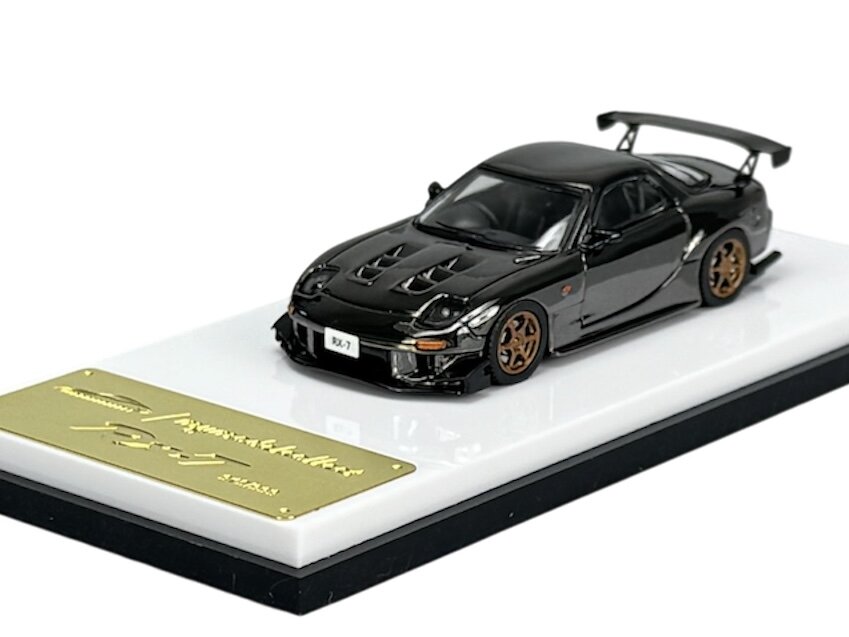 Model samochodu w kolorze ciemnym z brązowymi felgami, umieszczony na białej podstawce z metalową tabliczką. Na tabliczce widnieje napis \'RX-7\'.