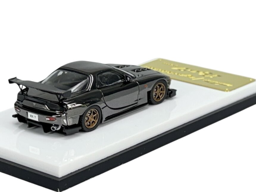 Model samochodu w kolorze gunmetal z brązowymi felgami, umieszczony na białej podstawce z napisem \'RX-7\' na tablicy rejestracyjnej.