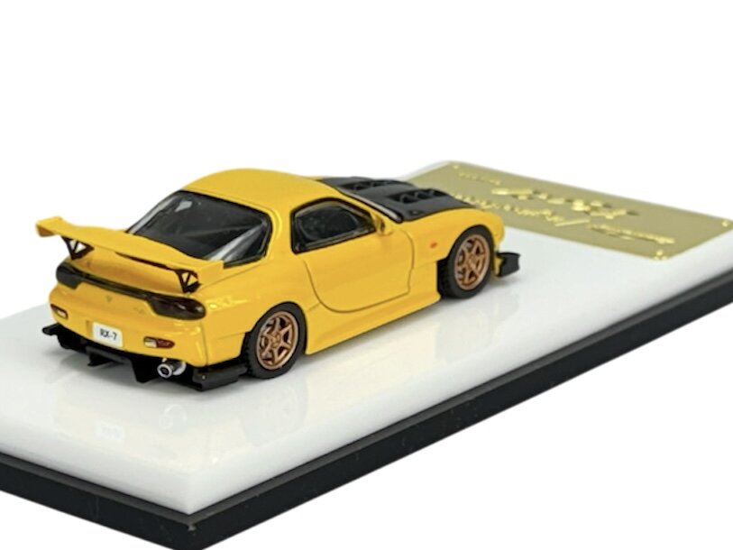Model samochodu w kolorze żółtym z czarnymi elementami, umieszczony na białej podstawce z czarną krawędzią. Na tablicy rejestracyjnej widnieje napis \'RX-7\'.