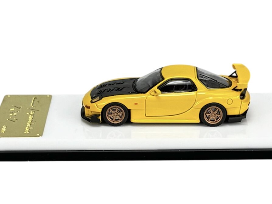 Model samochodu w skali 1:64 w kolorze żółtym z czarnymi detalami, umieszczony na białej podstawce z metalową tabliczką. Na tabliczce widoczny jest tekst, ale nie jest czytelny.