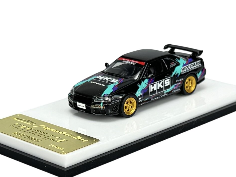 Model samochodu w skali 1:64 z czarnym nadwoziem, kolorowymi grafikami i złotymi felgami, umieszczony na białej podstawce z metalową tabliczką. Na samochodzie widoczne są napisy \'HKS\' i \'NISSAN\'.