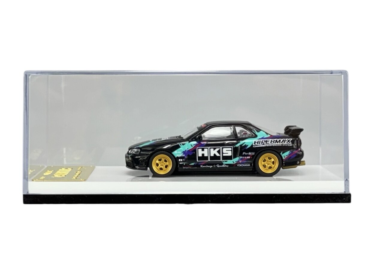 Model samochodu w skali 1:64 umieszczony w przezroczystej gablocie. Na boku pojazdu widoczne są napisy \'HKS\' i \'HIPERMAX\', a koła są w kolorze żółtym.