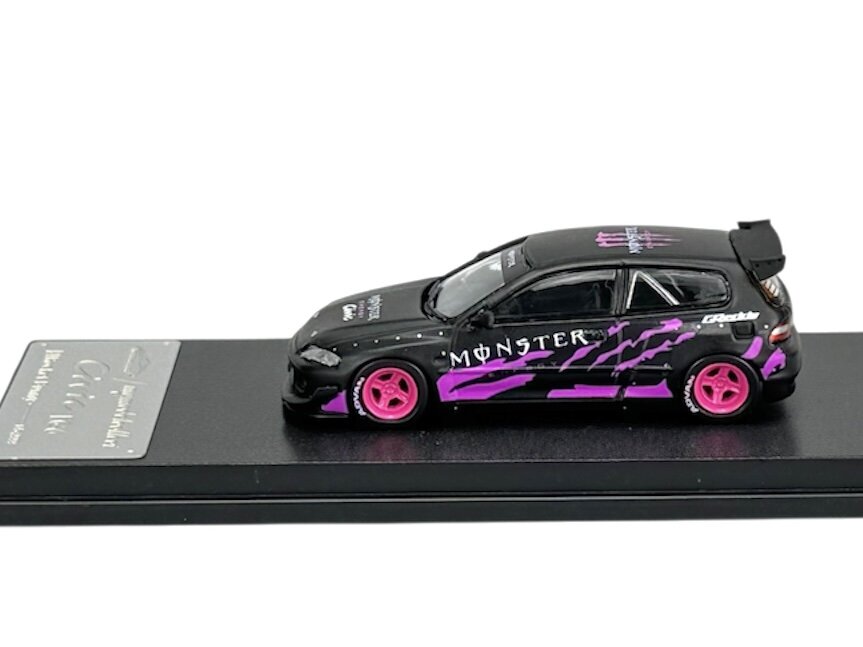 Model samochodu w skali 1:64 w kolorze czarno-fioletowym z różowymi felgami, umieszczony na czarnej podstawce. Na boku pojazdu widoczny jest napis \'MONSTER\'.
