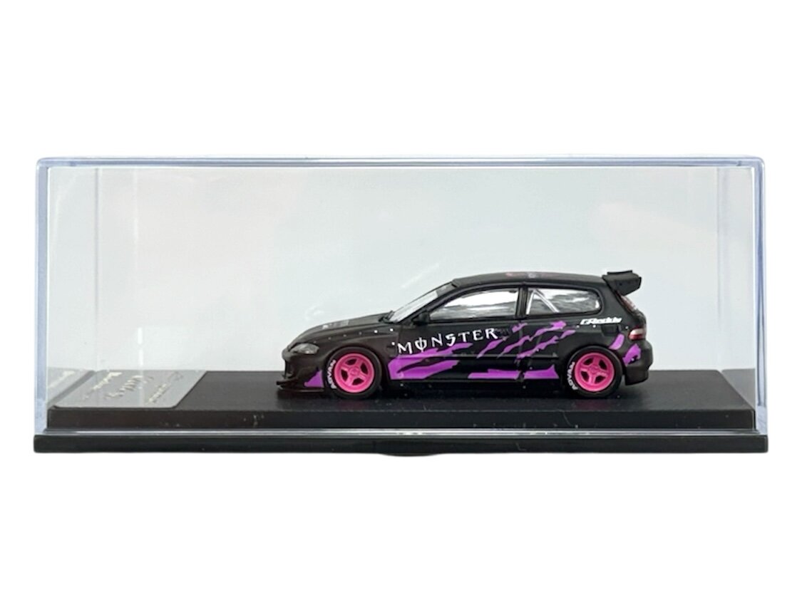 Model samochodu w skali 1:64 w kolorach czarnym i fioletowym z różowymi felgami, umieszczony w przezroczystej gablocie. Na boku pojazdu widoczny jest napis \'MONSTER\'.