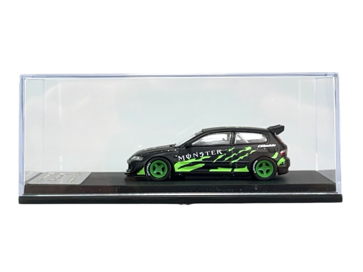 Model samochodu w skali 1:64 w kolorze czarnym z zielonymi akcentami i napisem \'Monster\' na boku, umieszczony w przezroczystej gablocie.