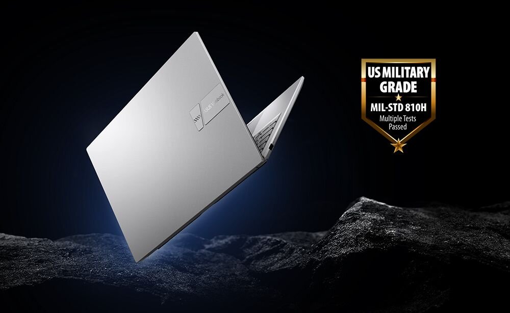 Srebrny laptop z logo producenta na pokrywie, uniesiony nad powierzchnią przypominającą skały. Obok widoczny jest emblemat z napisem \'US MILITARY GRADE MIL-STD 810H Multiple Tests Passed\'.