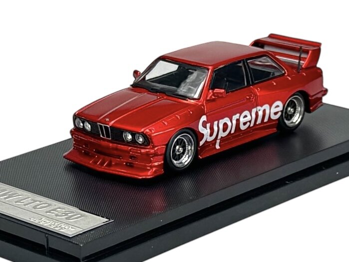 Czerwony model samochodu z napisem \'Supreme\' na boku, umieszczony na czarnej podstawce z napisem \'BMW E30\'.