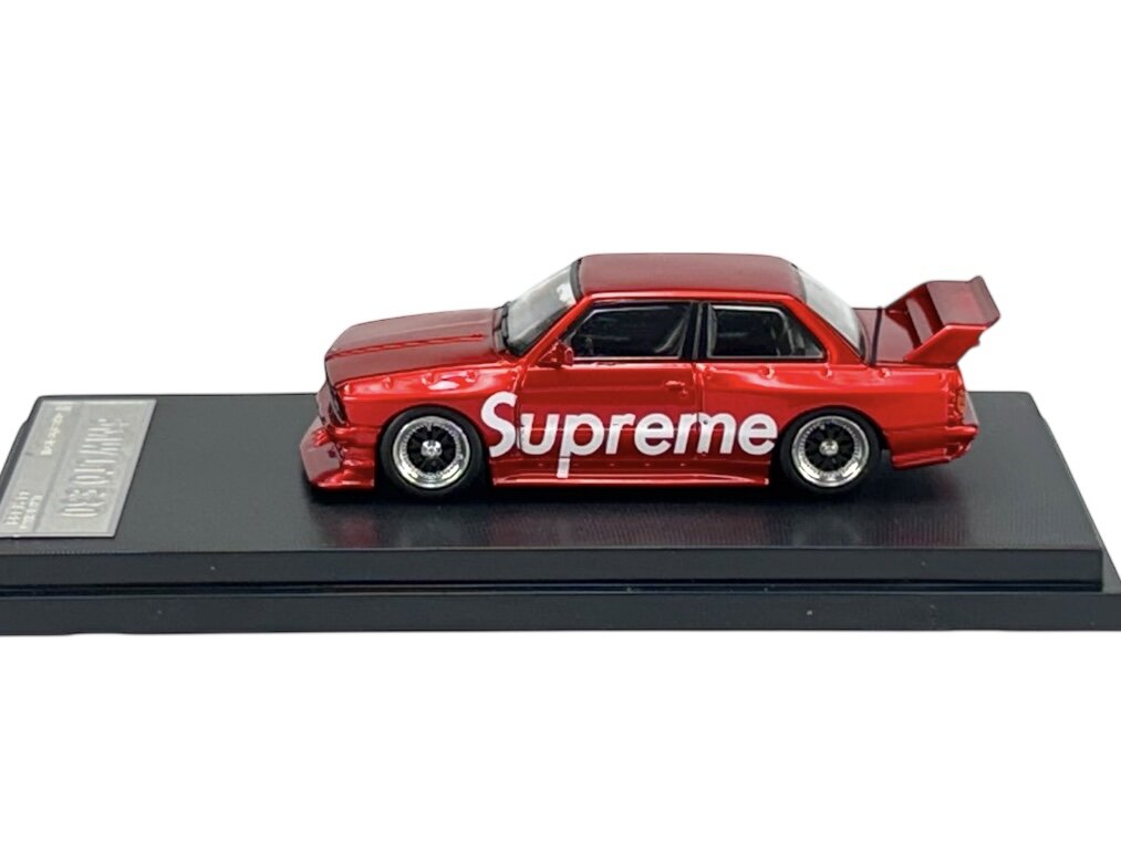 Czerwony model samochodu z napisem \'Supreme\' na boku, umieszczony na czarnej podstawce. Samochód ma duży tylny spojler i szczegółowe felgi.