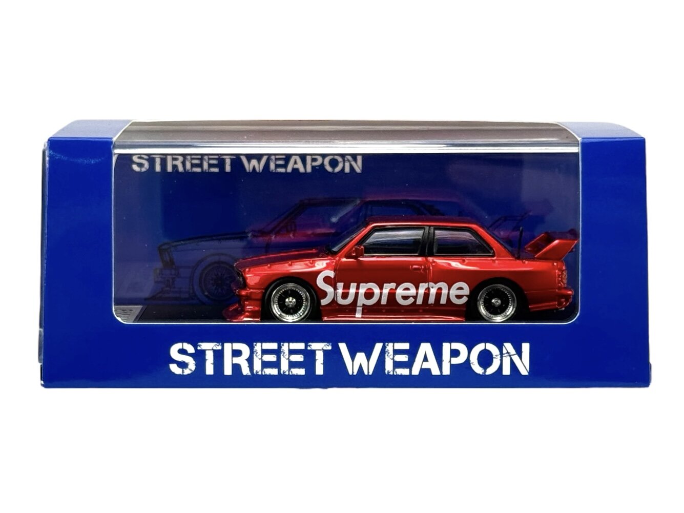 Czerwony model samochodu z napisem \'Supreme\' umieszczony w niebieskim pudełku z przezroczystym frontem. Na pudełku widnieje napis \'STREET WEAPON\'.