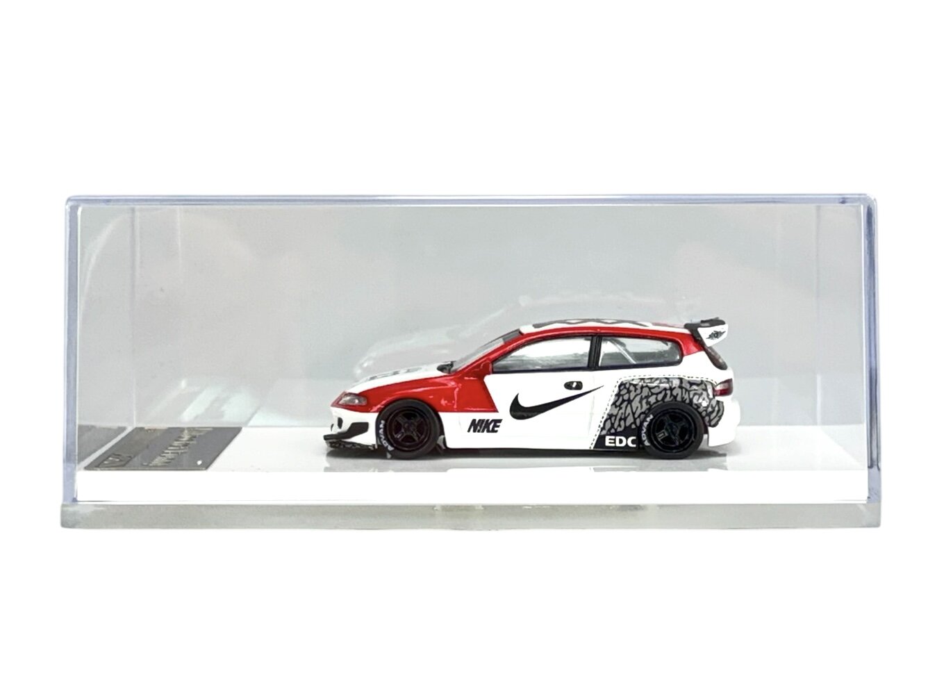 Model samochodu w skali 1:64 w kolorze biało-czerwonym z czarnymi detalami, umieszczony w przezroczystej gablocie. Na boku pojazdu widoczne są napisy \'NIKE\' i \'EDC\'.